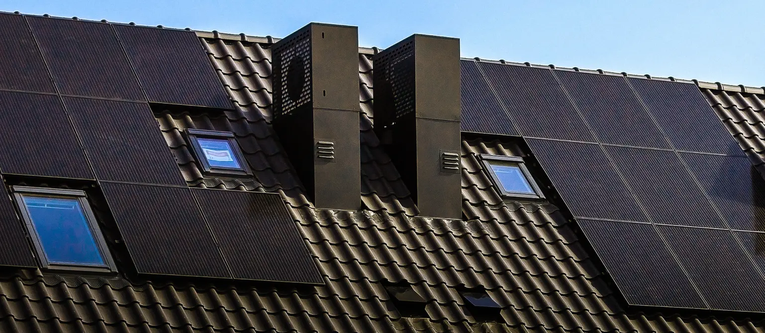 Ein dunkles Ziegeldach, auf dem schwarze Solarpaneele montiert sind. Daneben Schornsteine.