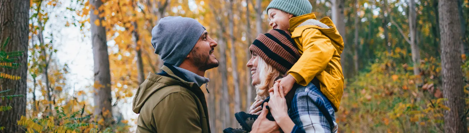 Nette dreiköpfige Familie beim Herbstspaziergang im Wald. Das Kind sitzt auf den Schultern der Mutter.