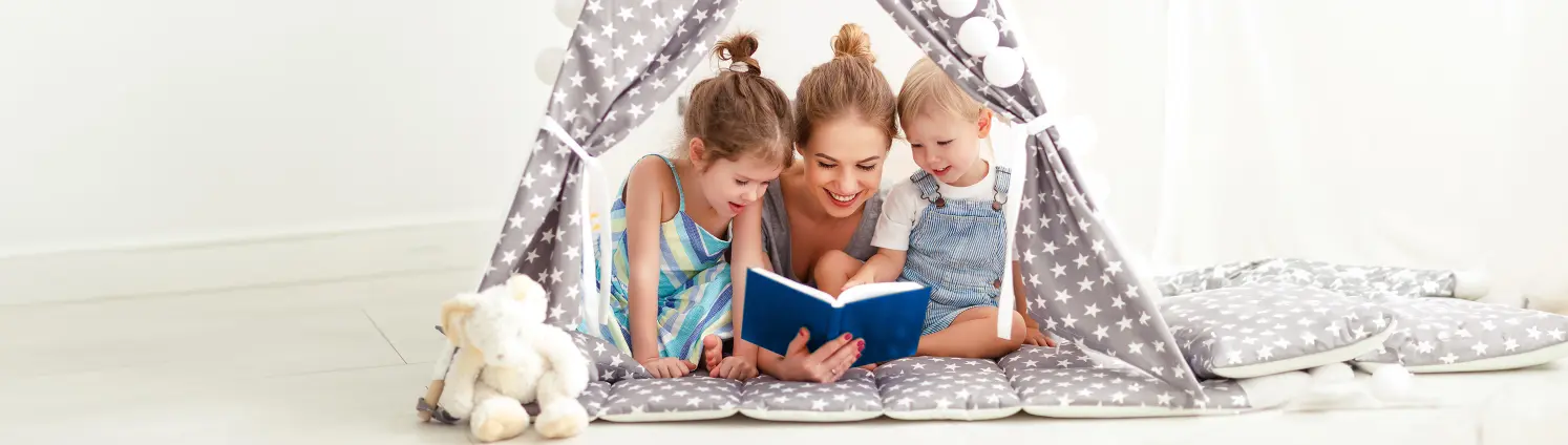Eine Frau liegt mit zwei Kindern in einem Spielzeugzelt und liest aus einem blauen Buch vor.