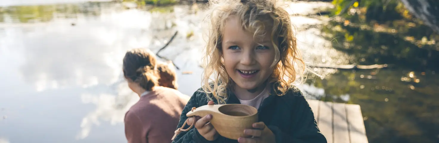 Ein Kind mit Becher in der Hand steht lächelnd auf einem Steg an einem See