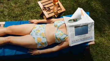 Eine Frausonnt sich in einem weiß-blau gestreiften Bikini mit Zitronen-Muster auf einer Liege im Garten und hat eine Zeitung über dem Gesicht.