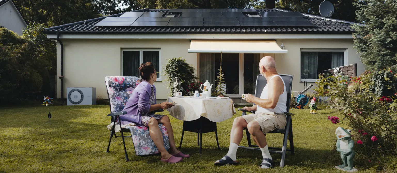 Älteres Pärchen sitzt im Garten und schaut nach hinten auf ihr Haus mit Photovoltaik