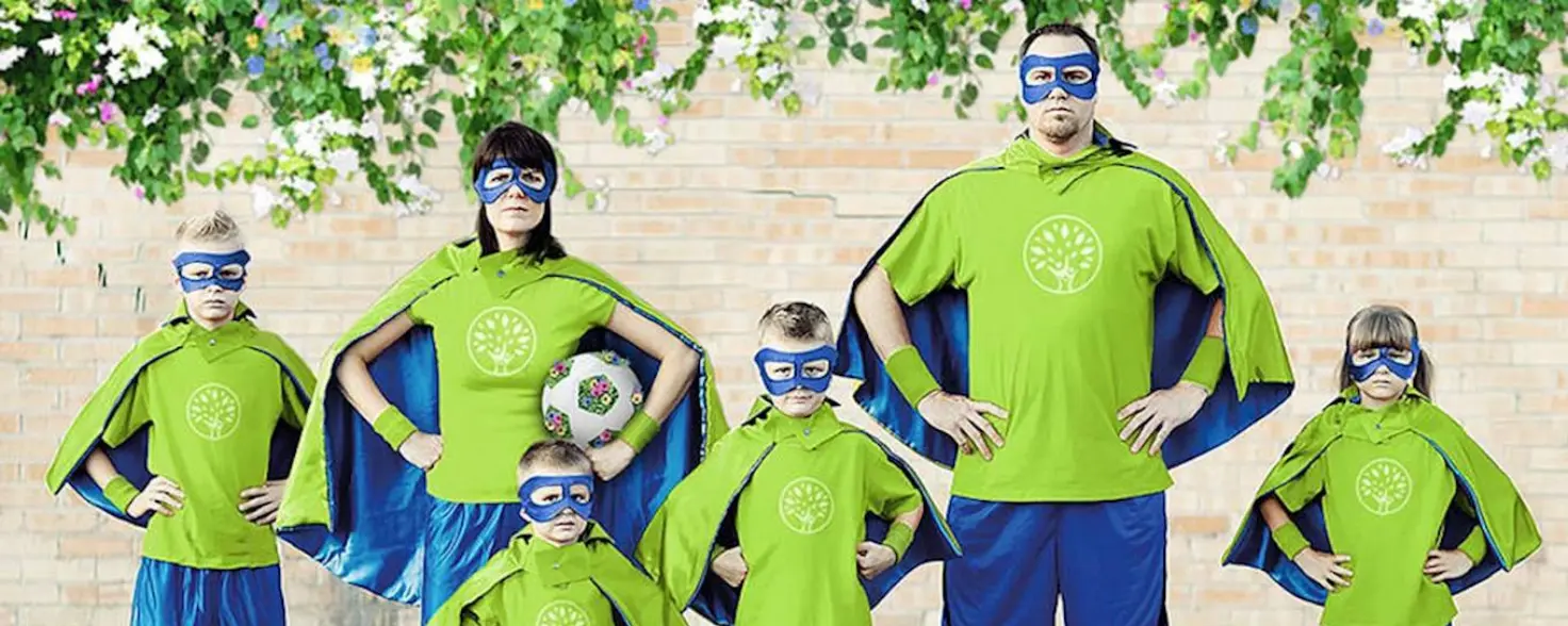 Eine Familie mit vier Kindern trägt blau-grüne Superhelden-Outfits. Die Mutter hat einen Fußball unter den Arm geklemmt.
