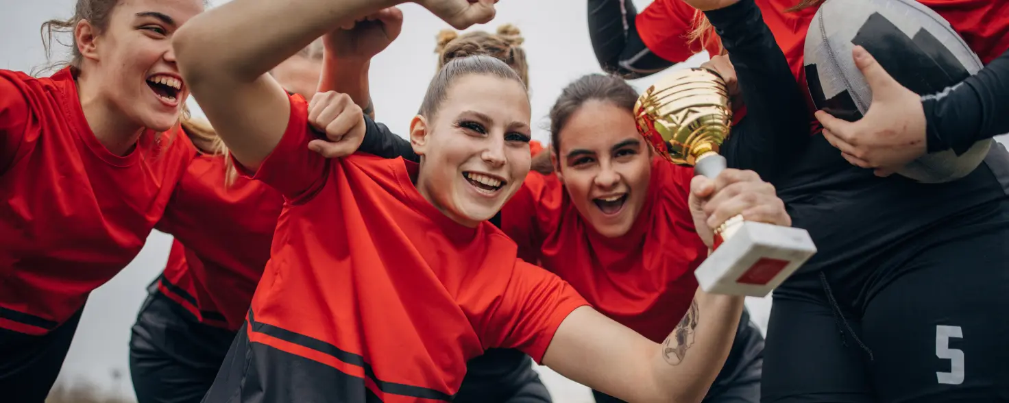 Sportlerinnen in rot-schwarzen Rugby-Trikots jubeln gemeinsam mit einem Siegerpokal auf dem Spielfeld.