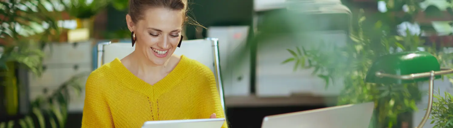 Eine Frau im gelben Pullover sitzt vor dem Laptop und hält ein Tablet in der Hand.