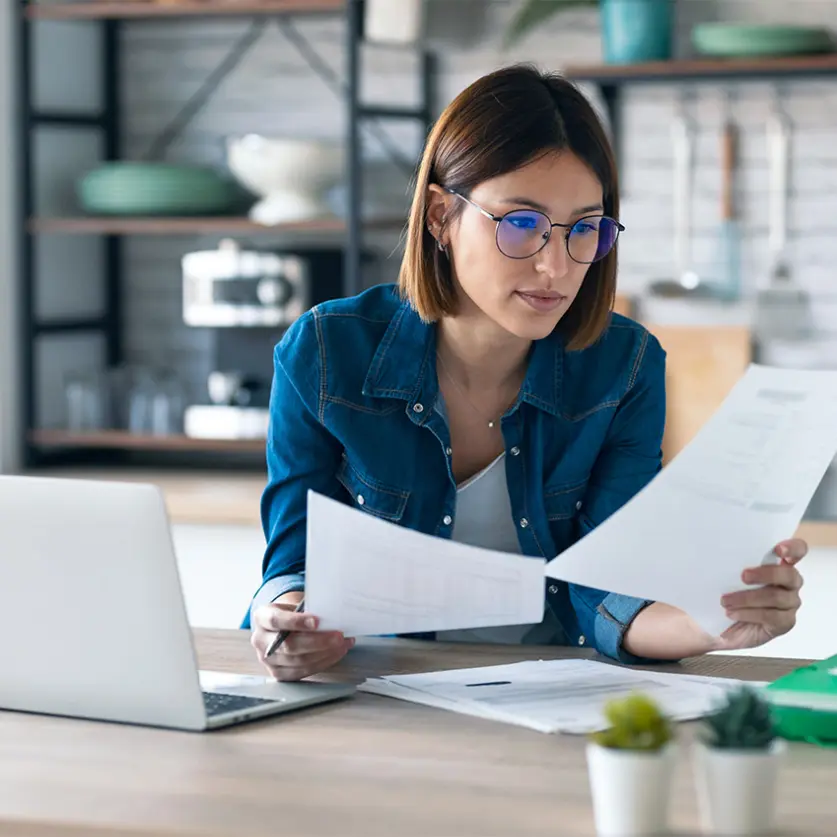 Eine Frau mit Brille sieht sich zwei ausgedruckte Seiten an. Sie sitzt an einem Schreibtisch mit Laptop. Bild mit KI erstellt.