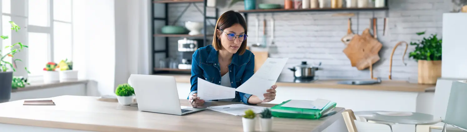 Eine Frau mit Brille sieht sich zwei ausgedruckte Seiten an. Sie sitzt an einem Schreibtisch mit Laptop.