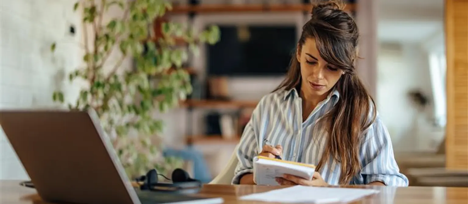 Eine Frau sitzt an einem großen Tisch vor ihrem Laptop und einem Notizheft in der Hand.