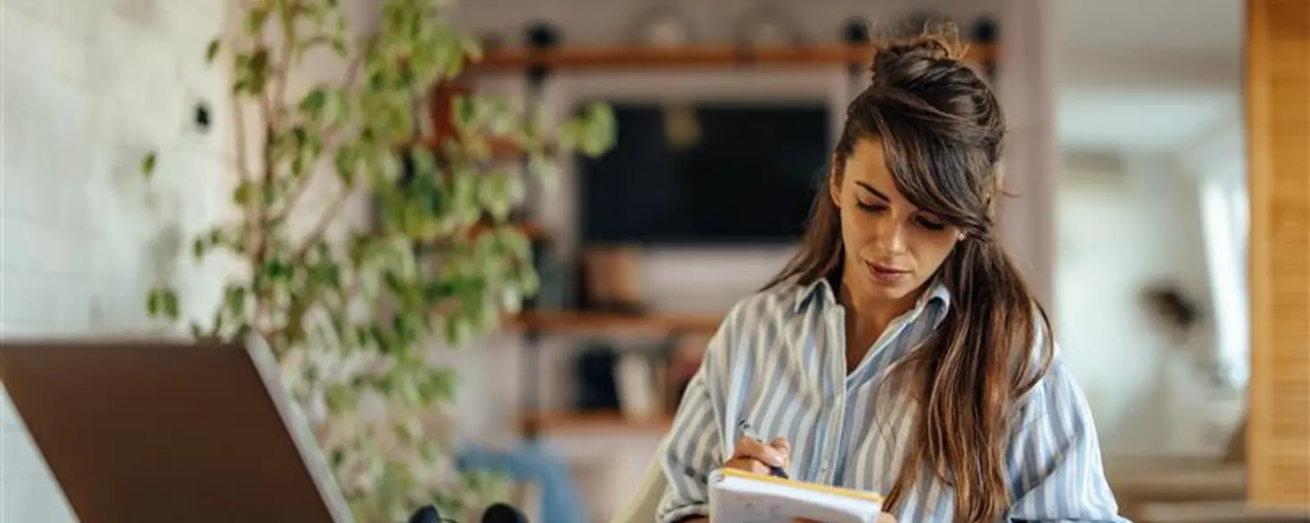 Eine Frau sitzt an einem großen Tisch vor ihrem Laptop und einem Notizheft in der Hand.