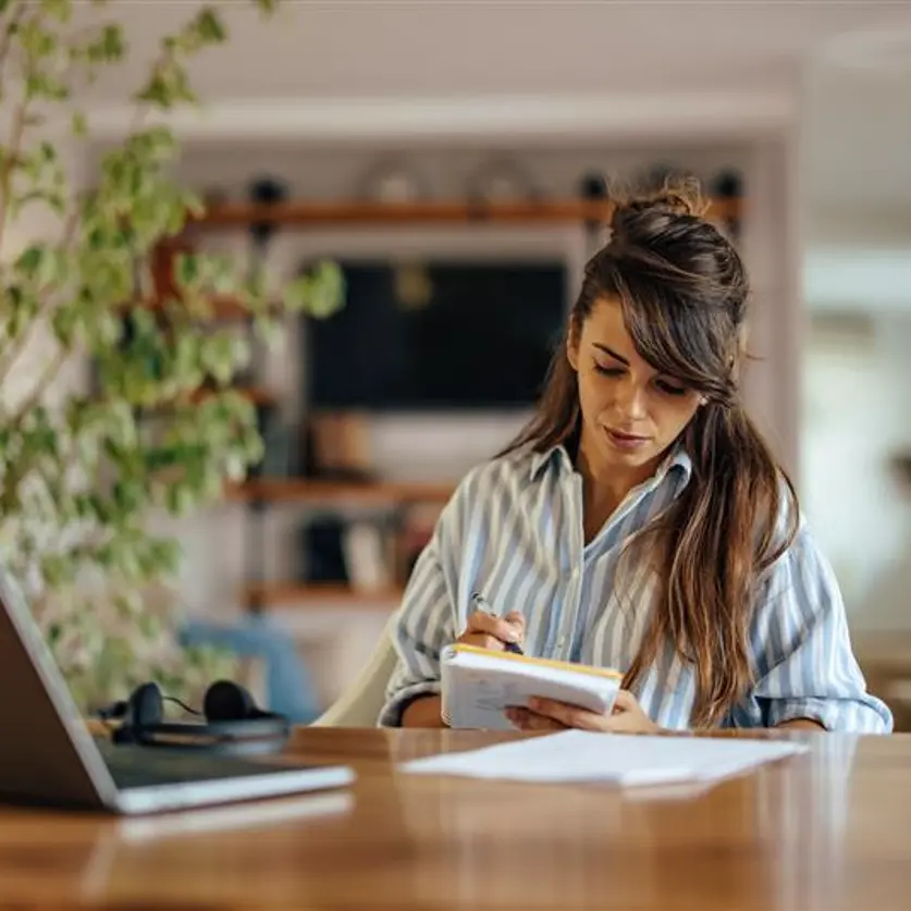 Eine Frau sitzt an einem großen Tisch vor ihrem Laptop und einem Notizheft in der Hand.