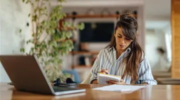 Eine Frau sitzt an einem großen Tisch vor ihrem Laptop und einem Notizheft in der Hand.