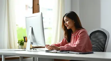 Eine Frau sitzt am Fenster vor ihrem silbernen Computerbildschirm und tippt auf der Tastatur.