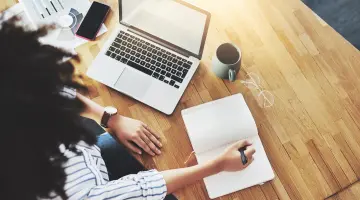 Sicht von oben: Eine Person arbeitet am Laptop und macht sich dabei Notizen in ein kleines Buch.