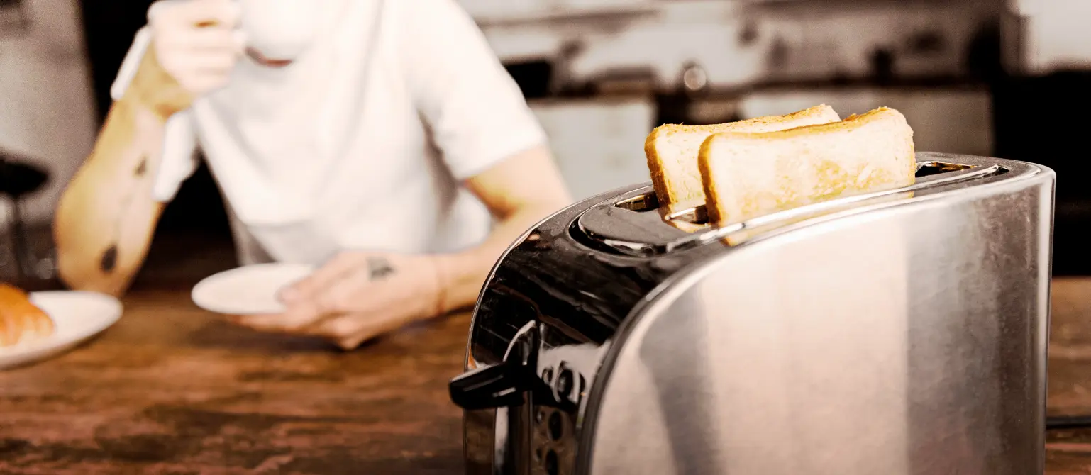 Im Vordergrund eine silberner Toaster und im Hintergrund ein Mann in einem weißen T-Shirt der aus einer Tasse trinkt