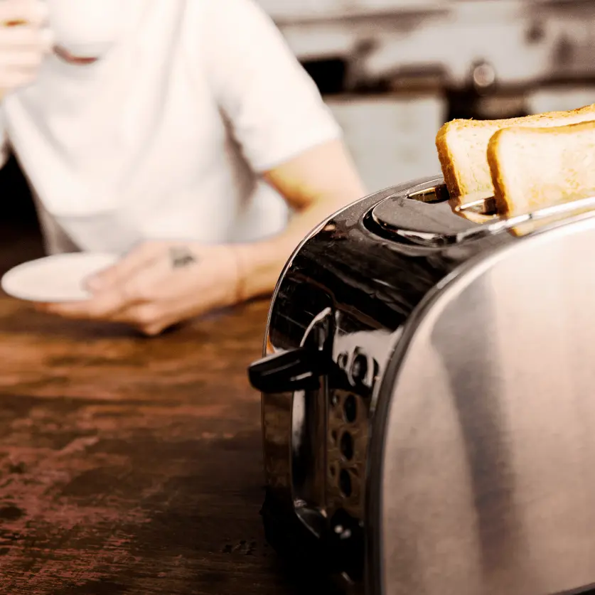 Im Vordergrund eine silberner Toaster und im Hintergrund ein Mann in einem weißen T-Shirt der aus einer Tasse trinkt