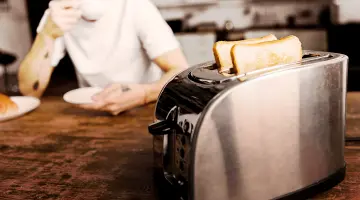 Im Vordergrund eine silberner Toaster und im Hintergrund ein Mann in einem weißen T-Shirt der aus einer Tasse trinkt