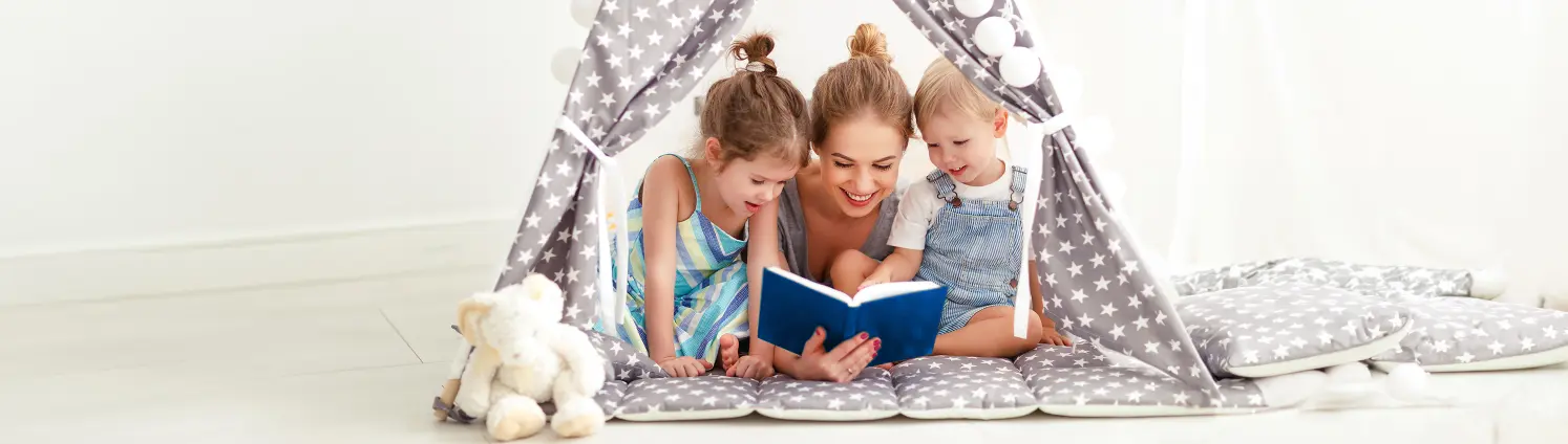 Eine Mutter liegt mit ihren zwei Kindern in einem Spielzeugzelt und liest aus einem blauen Buch vor.