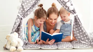 Eine Mutter liegt mit ihren zwei Kindern in einem Spielzeugzelt und liest aus einem blauen Buch vor.
