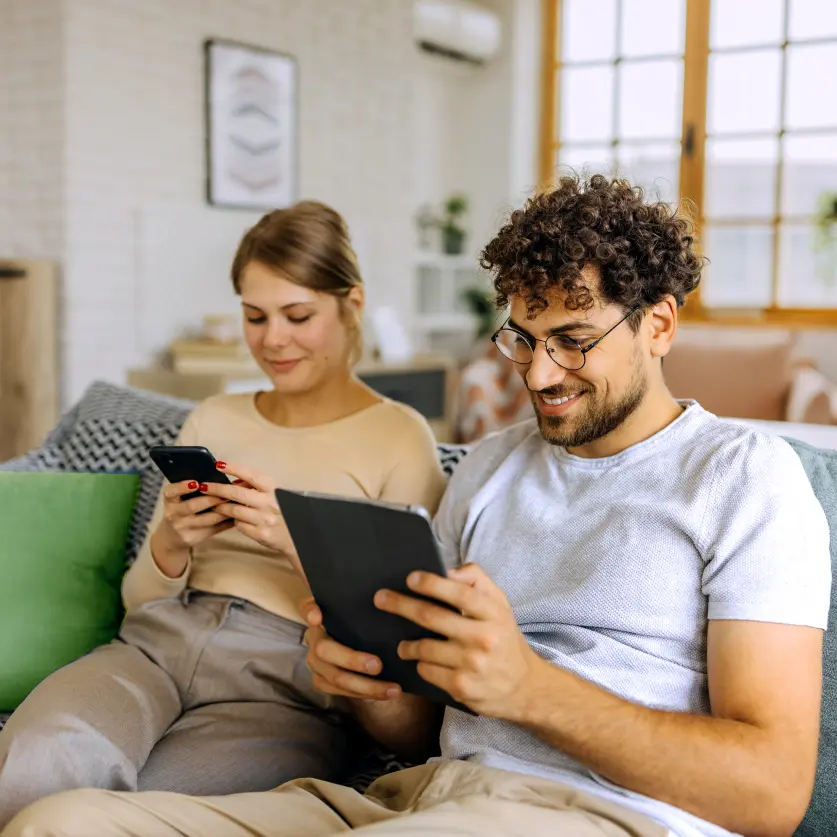 Gasanbieter wechseln: Eine Frau mit Pferdeschwanz und ein lockiger Mann sitzen im Wohnzimmer auf dem Sofa und surfen auf Tablet-PC und Handy.