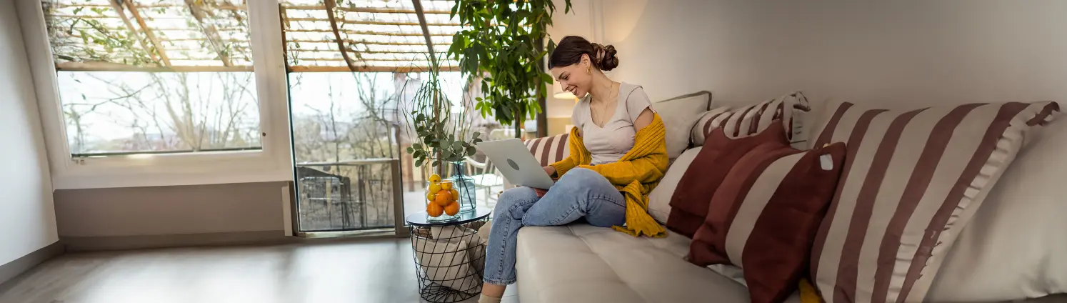 Frau sitzt im Wohnzimmer auf einer Couch mit ihrem Laptop auf den Knien.
