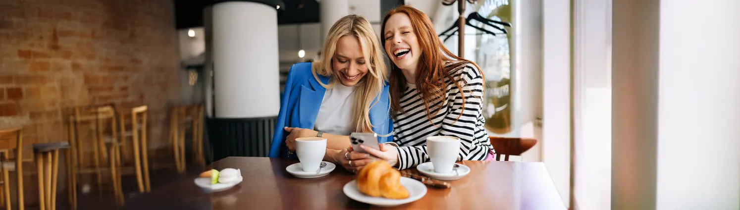 Zwei Frauen sitzen an einem Tisch mit Kaffeetassen und Gebäck, schauen auf ein Smartphone und lachen.