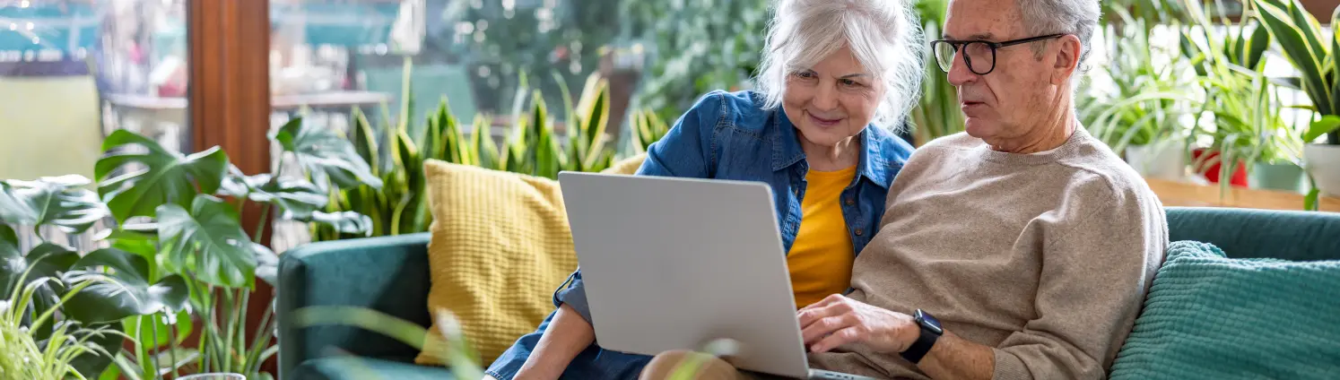 Seniorenpaar mit Laptop sitzt zu Hause auf dem Sofa im Wohnzimmer.
