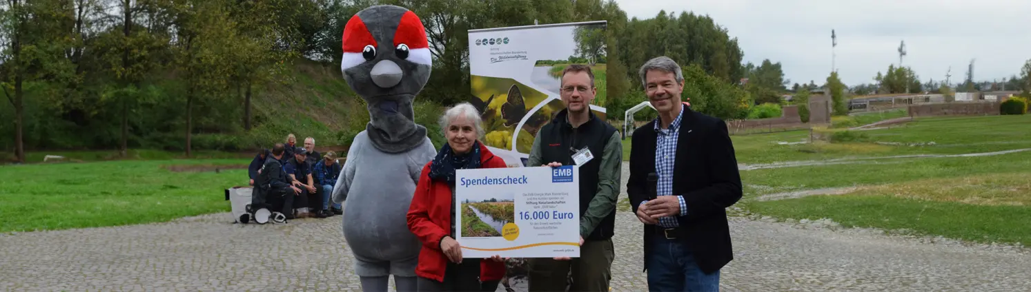 Ein Maskottchen und mehrere Personen halten einen Spendenscheck über 16.000 Euro. Im Hintergrund grüne Landschaft.