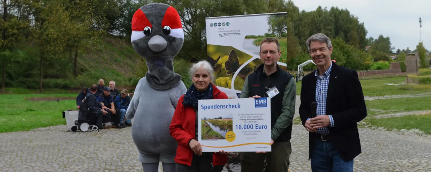 Ein Maskottchen und mehrere Personen halten einen Spendenscheck über 16.000 Euro. Im Hintergrund grüne Landschaft.