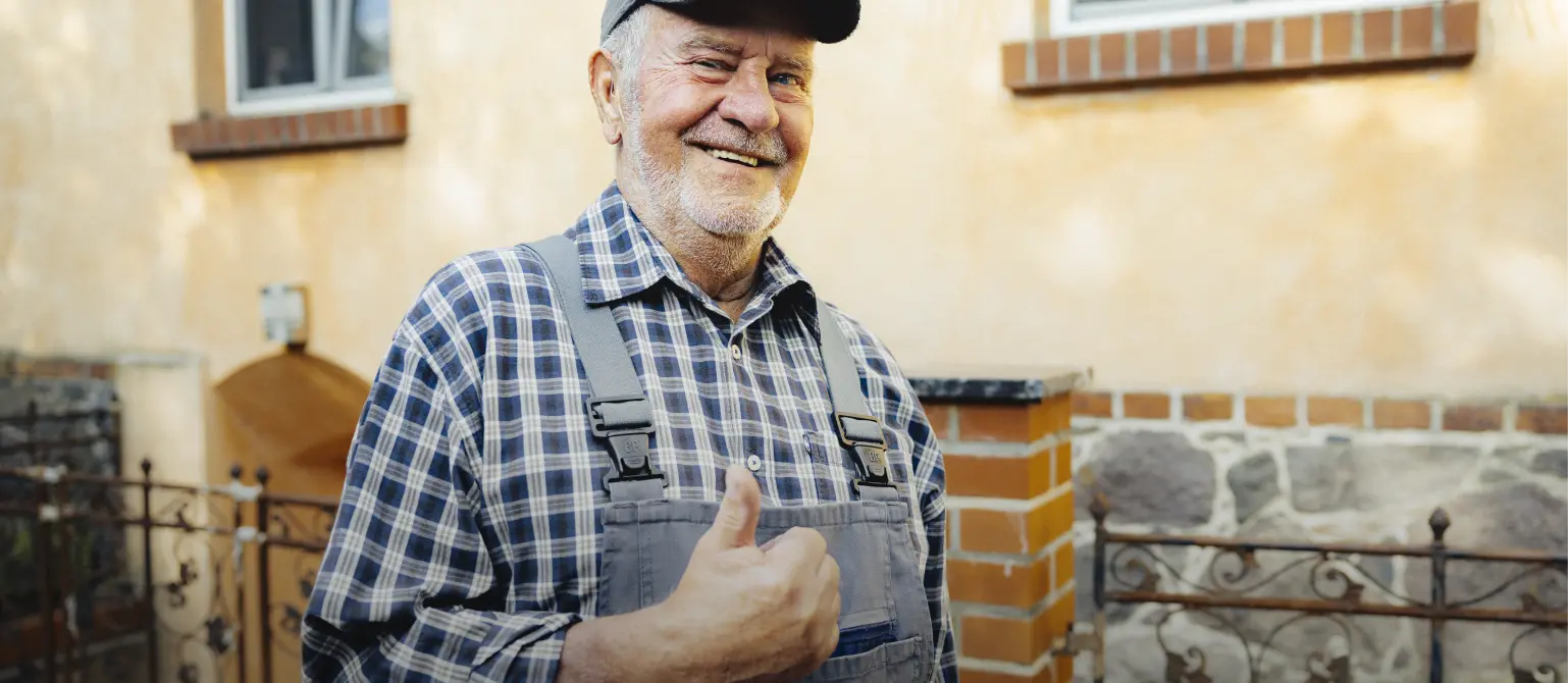 Ein älterer Handwerker in Arbeitskleidung und Basecap hebt augenzwinkernd den Daumen.