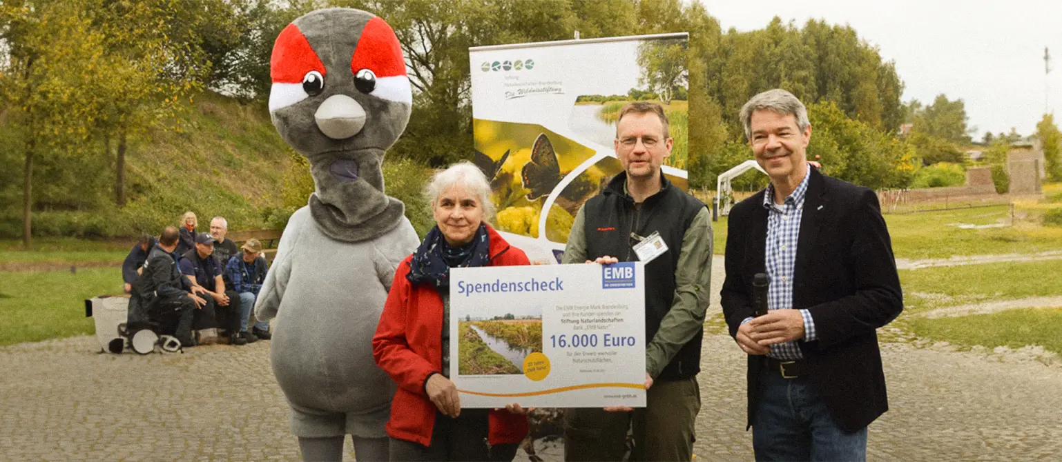 Ein Maskottchen und mehrere Personen halten einen Spendenscheck über 16.000 Euro. Im Hintergrund grüne Landschaft.