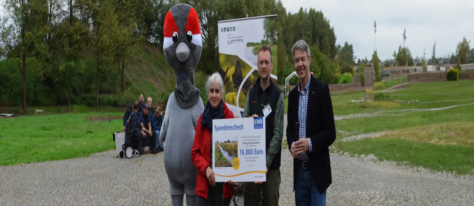 Ein Maskottchen und mehrere Personen halten einen Spendenscheck über 16.000 Euro. Im Hintergrund grüne Landschaft.