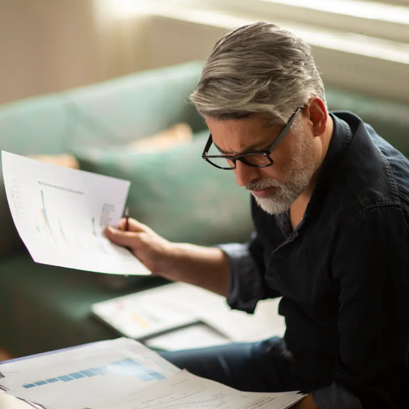 Älterer Mann mit Brille sitzt auf dem Sofa und studiert die Seiten einer ausgedruckten Rechnung.