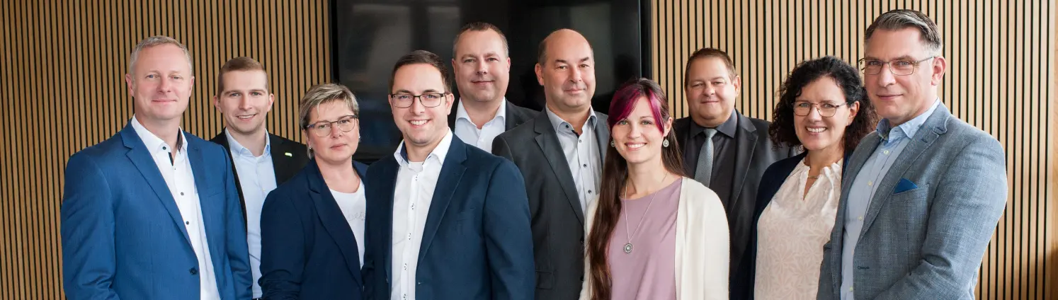 Mehrere Menschen vor einer Holzwand in schicken Business-Outfits schauen in die Kamera.