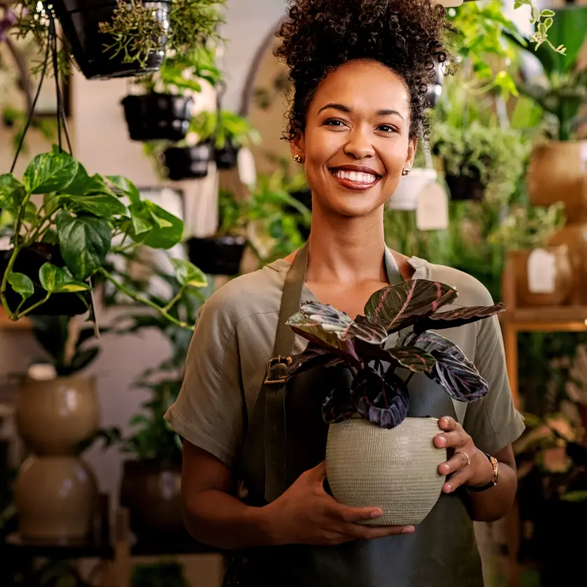 Eine lächelnde Frau mit lockigem Haar hält eine grüne Zimmerpflanze in einem Blumenladen.