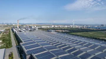 Große Industriehallen mit Photovoltaik-Anlagen auf dem Dach. Im Hintergrund Windkraftanlagen vor blauem Himmel.