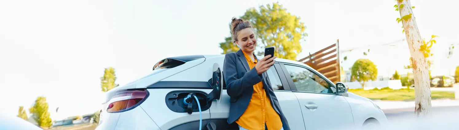 Eine Frau lehnt an einem weißen Auto, das gerade geladen wird und schaut lächelnd auf ihr Handy.