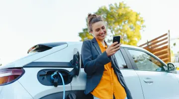 Eine Frau lehnt an einem weißen Auto, das gerade geladen wird und schaut lächelnd auf ihr Handy.