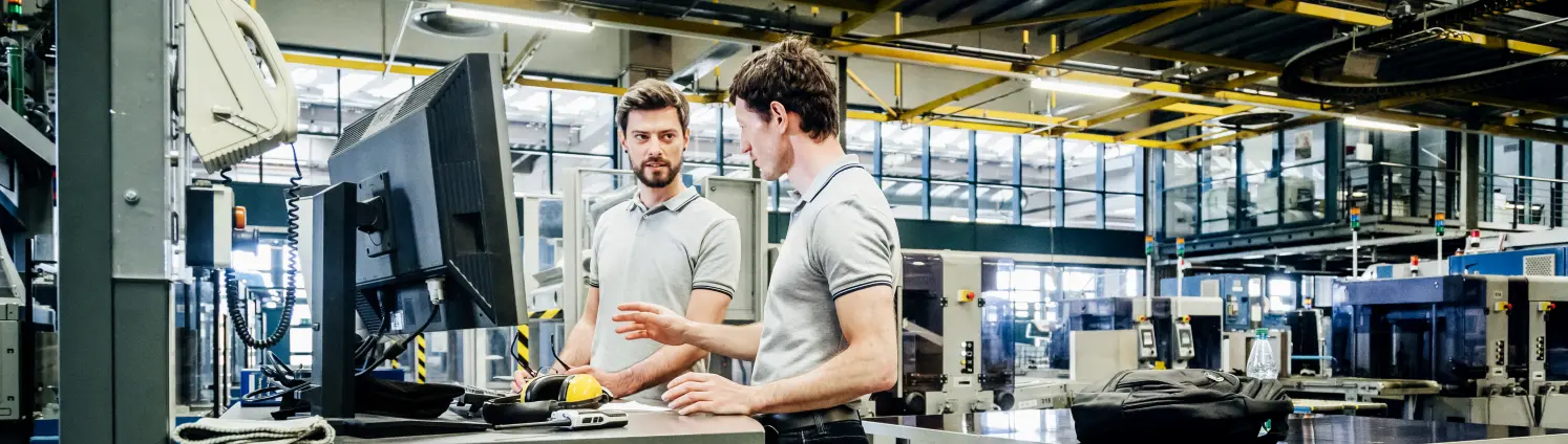 Zwei Männer stehen an einem PC in einer Fabrikhalle und diskutieren