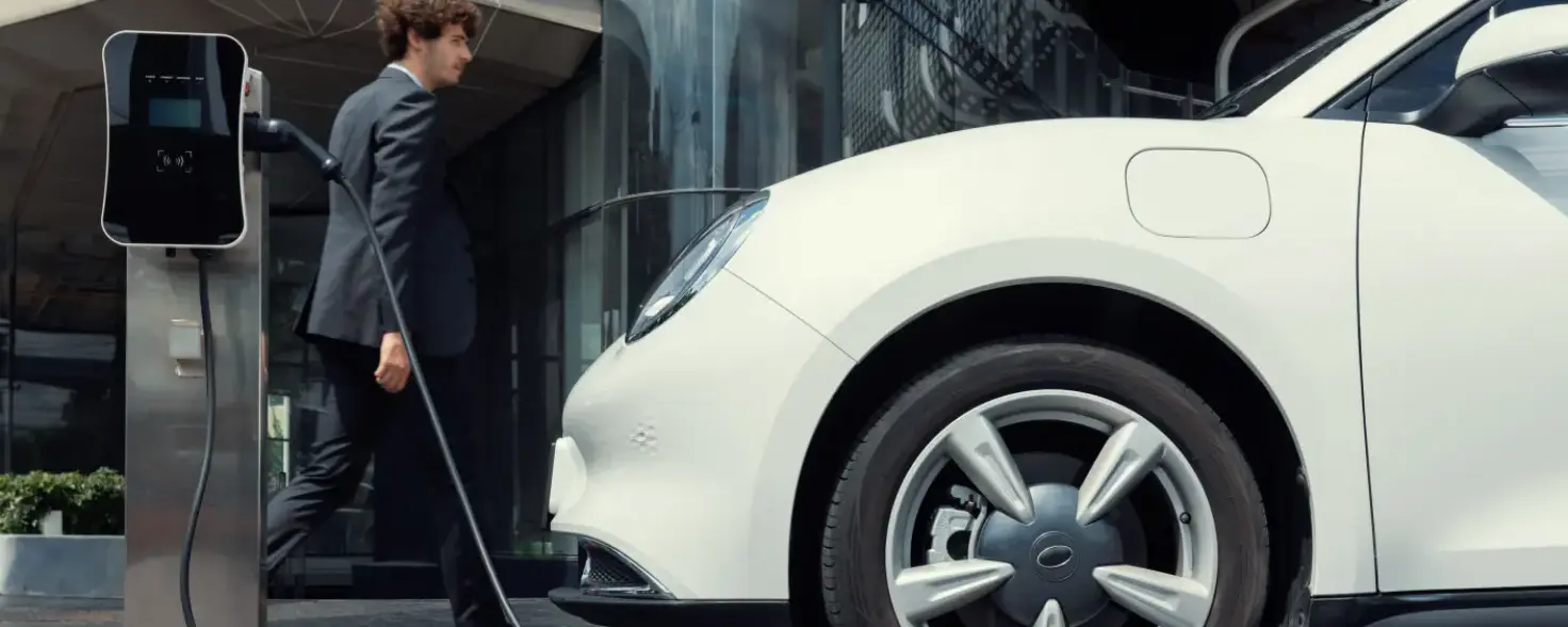 Links steht eine E-Auto-Ladesäule, rechts ein weißes Auto. Dazwischen geht ein Mann im Anzug entlang.