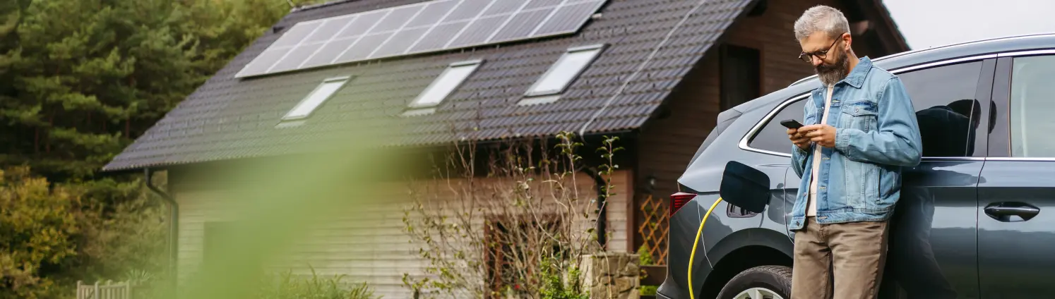 Ein Mann in Jeansjacke lädt sein Elektroauto vor einem Einfamilienhaus mit Photovoltaik-Anlage und blickt dabei auf sein Mobiltelefon.