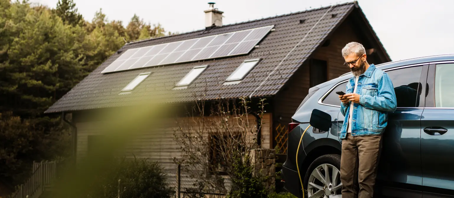 Ein Mann in Jeansjacke lädt sein Elektroauto vor einem Einfamilienhaus mit Photovoltaik-Anlage und blickt dabei auf sein Mobiltelefon.