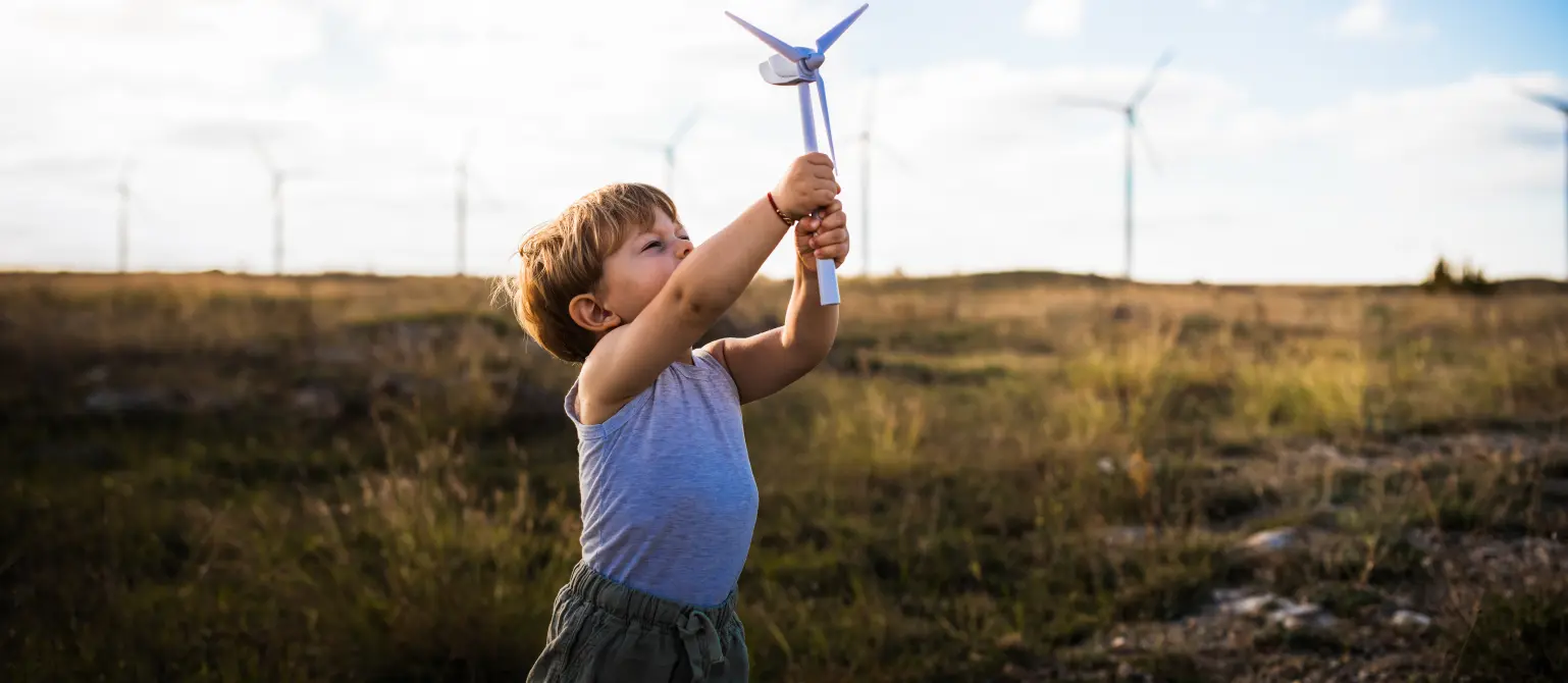 Landschaft mit Windkraftanlagen: Ein kleiner Junge reckt ein Modell eines Windrads in den blauen Himmel.