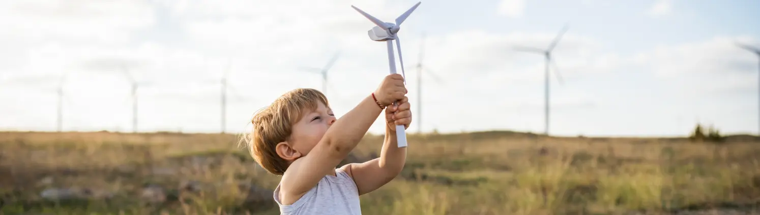 Landschaft mit Windkraftanlagen: Ein kleiner Junge reckt ein Modell eines Windrads in den blauen Himmel.
