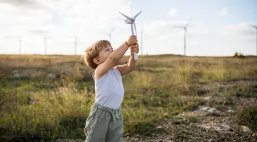 Landschaft mit Windkraftanlagen: Ein kleiner Junge reckt ein Modell eines Windrads in den blauen Himmel.