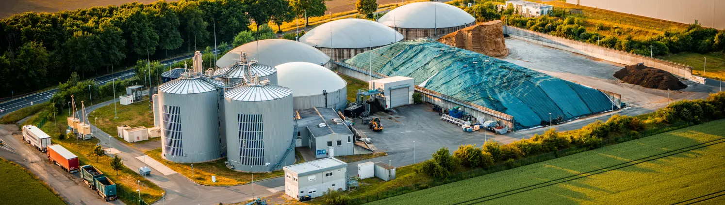 Luftaufnahme einer Biogasanlage mit Silos und Rohstofflager vor der eine Reihe von LKW wartet. Drumherum Felder und Bäume.