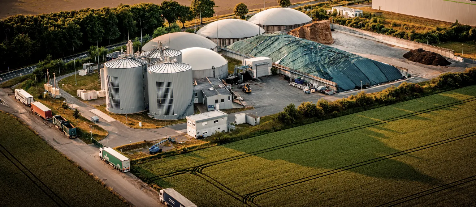 Luftaufnahme einer Biogasanlage mit Silos und Rohstofflager vor der eine Reihe von LKW wartet. Drumherum Felder und Bäume.