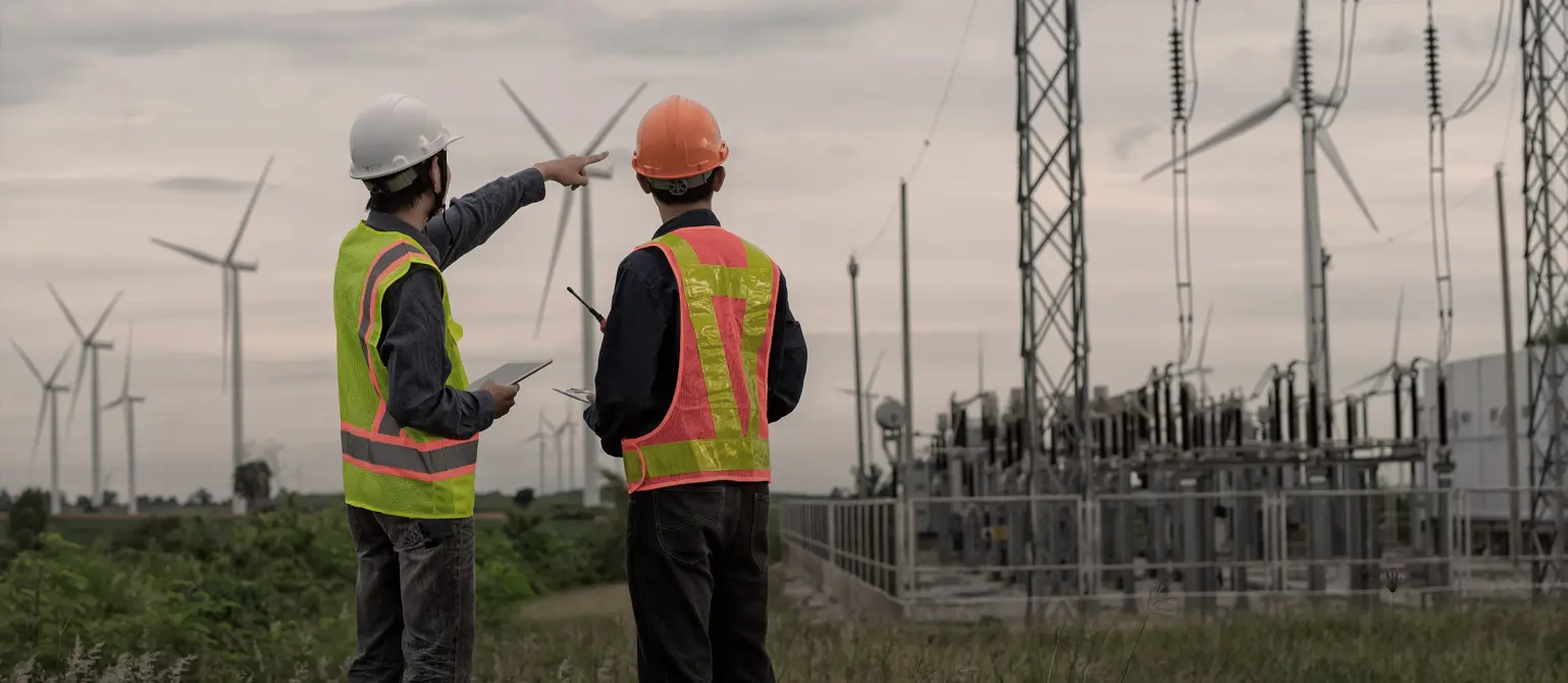 Zwei Ingenieure in Helmen und Sicherheitswesten prüfen elektrisches Umspannwerk und benachbarte Windkraftanlagen.