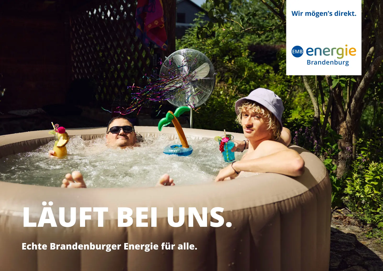 Zwei junge Männer mit Sonnenhut und Sonnenbrille lümmeln in einem aufblasbaren Whirlpool und trinken bunte Cocktails.