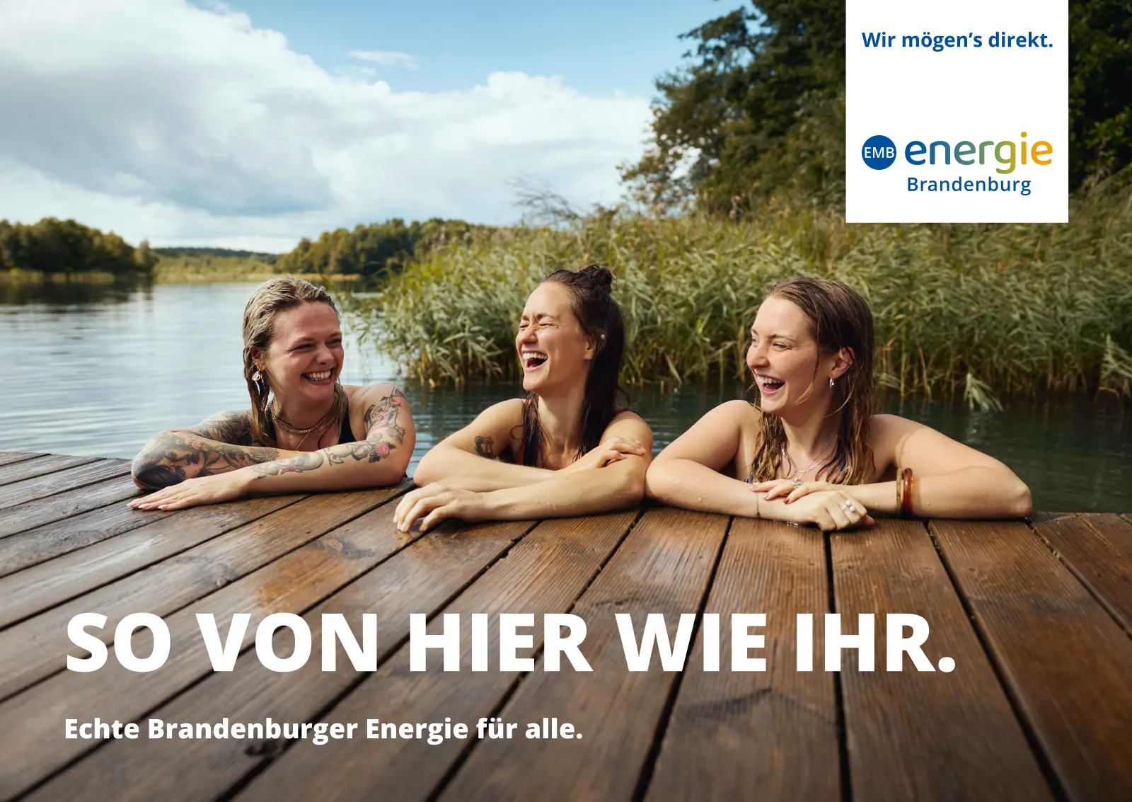 So von hier wie ihr: Drei junge Frauen im Wasser eines Sees halten sich an einem Holzsteg fest und lachen aus vollem Hals.