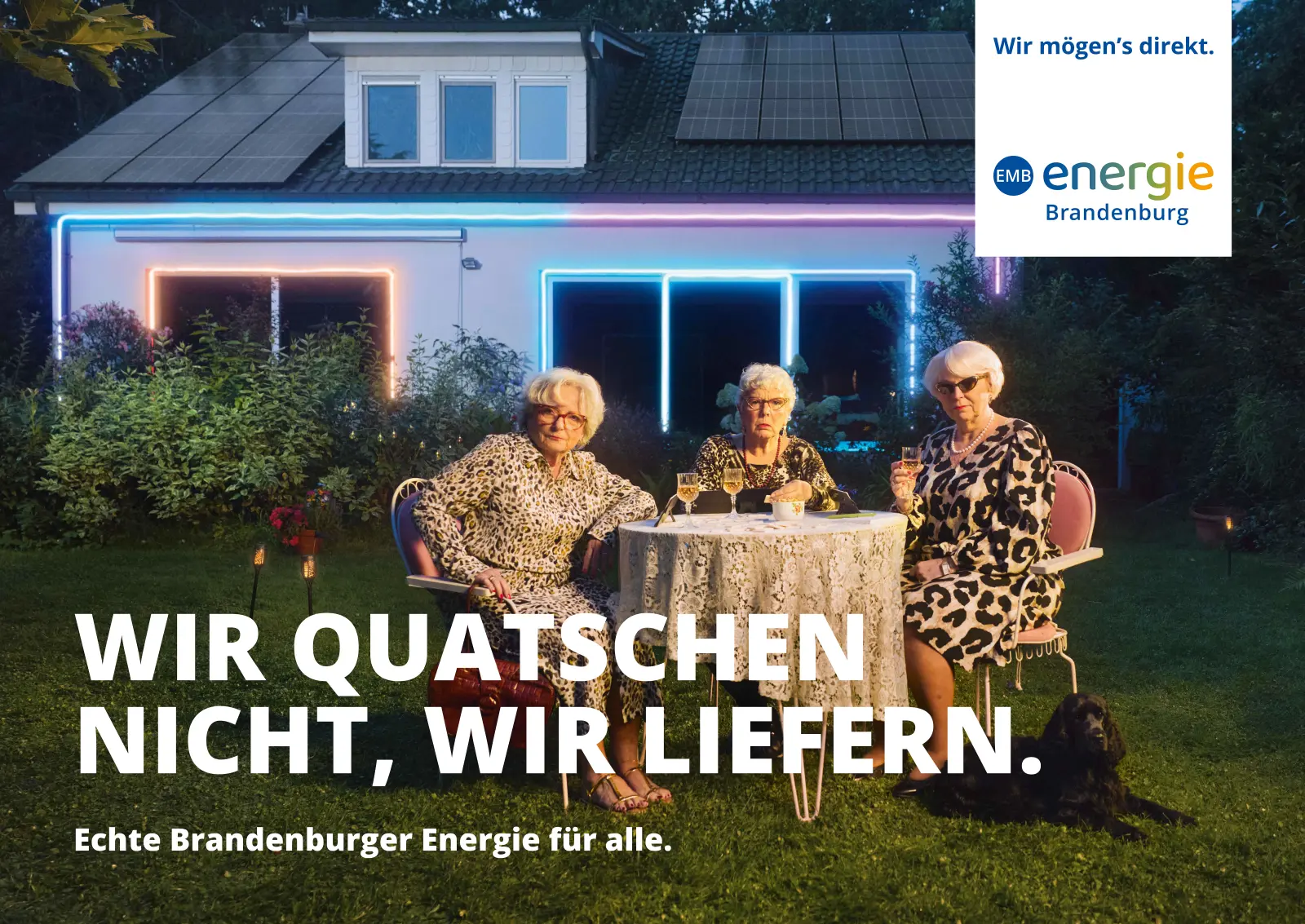 Drei ältere Frauen in gemusterten Kleidern spielen am Gartentisch Karten. Im Hintergrund ein Bungalow mit PV-Anlage auf dem Dach.